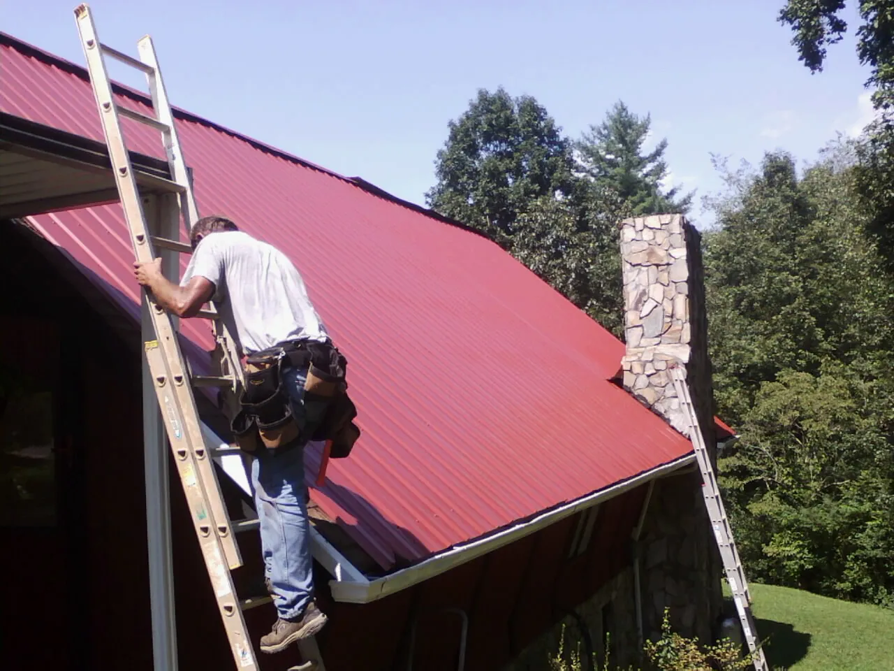 Roofer on a ladder beside a red metal roof and stone chimney — Soffit & Fascia Repair by Roofing Brothers in Nahant